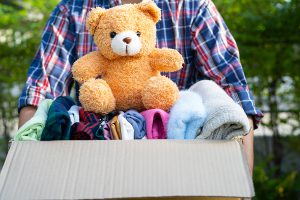 Someone holding a box full of clothes to donate, with a teddy bear sitting on top